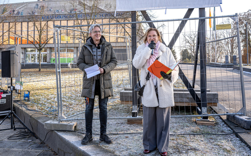 Zwei Personen, ein Mann und eine Frau, stehen vor einem Gebäude mit einem Stahlgerüst und sprechen in die Mikrofone. Sie sind in Mänteln gehüllt und halten dabei Papier in der Hand.