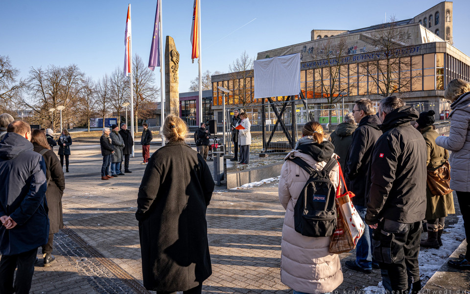 Eine Gruppe von Menschen steht vor einem Theatergebäude und betrachtet eine weiße Struktur mit einem Holzstatuen. Die Szene spielt an einem sonnigen Wintertag in einem öffentlichen Platz mit einer Pflasterstrecke.