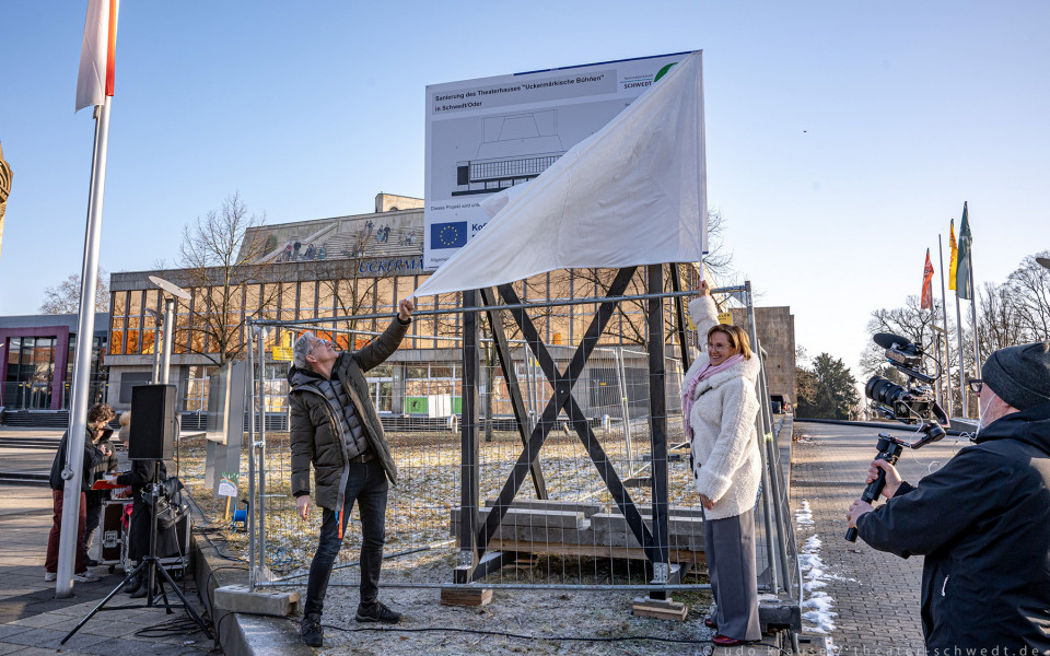 Auf dem Foto sehen zwei Frauen und eine Gruppe von Journalisten vor einem Baugerüst vor einer Theaterfassade. Ein großes weißes Banner mit Bauzeichnungen und Informationen hängt an dem Gerüst, während eine Person es abdeckt.