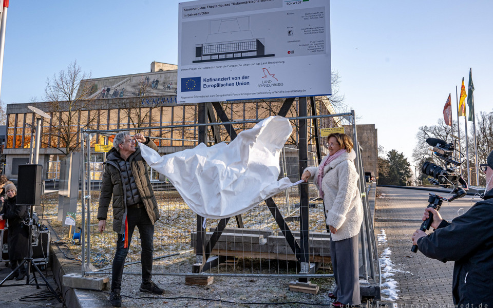 Auf dem Foto ist eine weiße Leinwand vor einem Theatergebäude ausgebreitet, während eine Gruppe von Menschen, darunter Journalisten und Fotografen, sie betrachtet. Hintergrund sind Menschen und ein Baum, während ein Projektlogo und ein Wappen des Europäischen Fonds für regionale Entwicklung zu sehen sind.