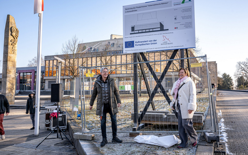 Auf einem Platz vor einem Theatergebäude stehen zwei Personen vor einer Bushaltestelle, während im Hintergrund Bauarbeiten an der Fassade zu sehen sind. Im Vordergrund sind verschiedene Kameras und weiteres Equipment zu erkennen.