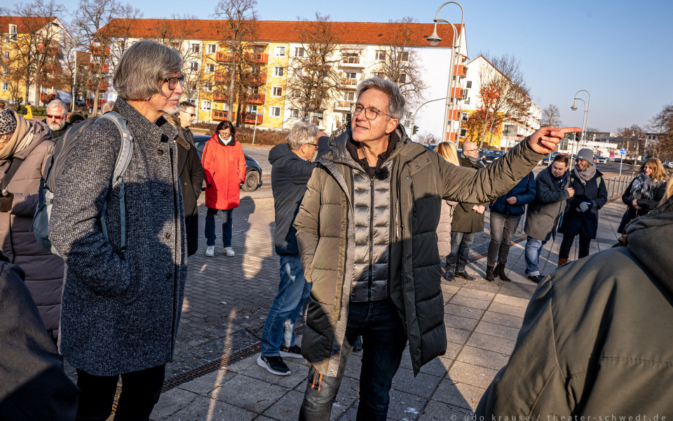 Eine Gruppe von Menschen, überwiegend ältere Damen und Herren, steht auf einer Promenade und scheint sich in einem Gespräch zu befinden. Ein Mann deutet mit dem Finger auf etwas in der Ferne, während die anderen Personen aufmerksam zuhören.