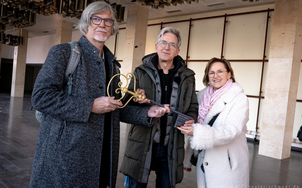 Drei Personen – ein Mann und zwei Frauen – stehen in einem Theaterflur und lächeln in die Kamera. Sie halten eine goldene Skulptur, die vermutlich mit dem Theater in Verbindung steht.