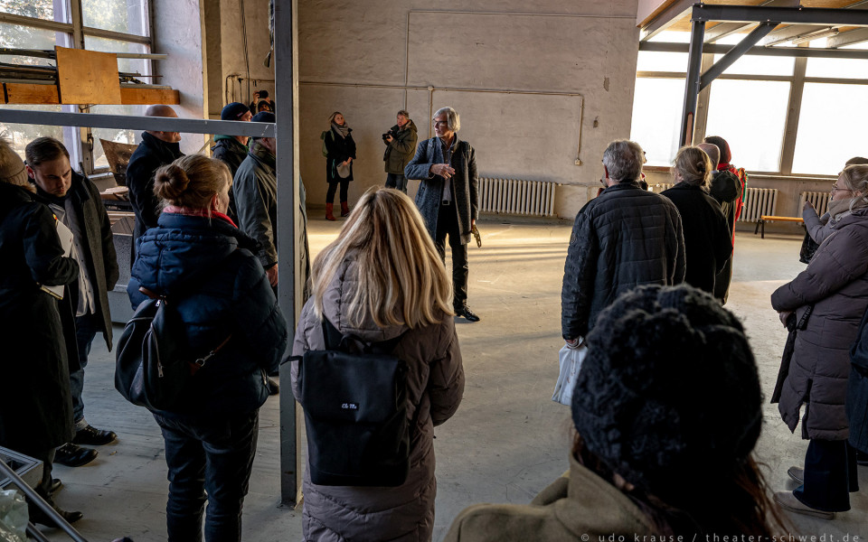 Es handelt sich um eine Gruppe von Menschen in einem großen, grauen Raum, vermutlich in einem Theater oder einer ähnlichen Veranstaltungshalle. Eine Person im Vordergrund spricht, während die anderen Zuschauer sind und vermutlich auf eine Vorstellung warten.