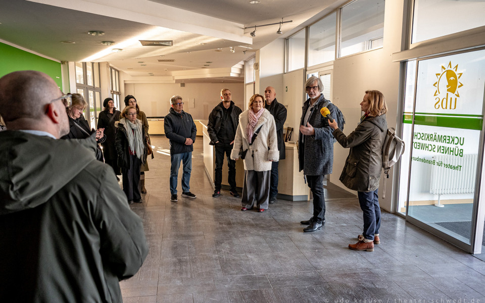 Auf dem Foto sind eine Gruppe von Menschen in einem modernen Theatergebäude zu sehen, während ein Reporter mit einem Mikrofon mit einer Frau spricht. Die Gruppe scheint bei einer Veranstaltung oder Pressekonferenz zu sein, wie die gedruckten Textzeilen auf der Tür zeigen.