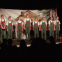 Mehrere Kinder stehen in identischen grünen Latzhosen, weißen T-Shirts und grauen Mützen auf einer Theaterbühne vor einem bemalten Bühnenbild mit Hausdach. Im Vordergrund sind die Silhouetten des Publikums zu sehen, während rechts ein Korb und links eine Krücke als Requisiten erkennbar sind.