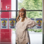 Ein Kind mit Brille, weißem Hemd und brauner Lederkappe steht in einem hellen Innenraum vor großen Fenstern. Im Hintergrund hängen mehrere Plakate für ein Theaterfest, während draußen grüne Bäume zu sehen sind.