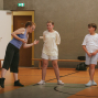 Eine Frau steht in einer Sporthalle und gestikuliert, während sie mit zwei Kindern spricht. Die Kinder tragen helle Sportkleidung und stehen auf dem Hallenboden vor einer grauen Wand und einer offenen Tür.