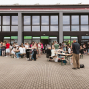 Vor einem modernen Gebäude mit großen Fenstern versammeln sich viele Menschen auf einem gepflasterten Platz. Einige sitzen an Biertischen und essen oder unterhalten sich, während andere in Gruppen vor den Eingängen stehen.