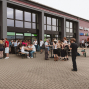 Vor einem modernen Gewerbegebäude mit großen Fenstern versammelt sich eine größere Gruppe von Menschen auf einem gepflasterten Platz. Einige sitzen an Biertischen, andere stehen in kleinen Gruppen zusammen, während im Hintergrund Lieferwagen und eine farbige Ladenbeschriftung zu sehen sind.