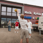Eine Gruppe junger Menschen spielt auf einem gepflasterten Platz vor einem Gebäude mit der Aufschrift „Maik Rybin Cars & Co“ mit einem gelb-schwarzen Ball. Rechts stehen ein weißer Lieferwagen und ein kleiner Transporter, während mehrere Personen dem Spiel zusehen.