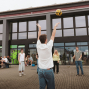 Eine Gruppe junger Menschen spielt auf einem gepflasterten Platz vor einem modernen Gebäude mit großen Fenstern Volleyball. In der Mitte wirft ein Mann in weißem T-Shirt den Ball hoch, während die anderen Spielerinnen und Spieler aufmerksam zusehen.