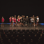 Eine Gruppe von Jugendlichen und einigen Erwachsenen steht auf einer dunklen Theaterbühne und scheint zu proben oder Anweisungen zu erhalten. Im Vordergrund sind leere Zuschauerreihen zu sehen, während die Bühne nur schwach beleuchtet ist.