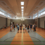 In einer großen Sporthalle steht eine Gruppe von Kindern und Jugendlichen im Kreis und hört einer Betreuerin zu. An den Wänden sind Bänke, hohe Fenster und ein Basketballkorb zu sehen.