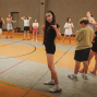 In einer Sporthalle stehen mehrere Kinder und Jugendliche verteilt auf dem Spielfeld, einige barfuß oder in Sportsachen. Im Vordergrund posiert ein Mädchen in schwarzer Kleidung mit erhobenen Fingern und herausgestreckter Zunge, während die anderen sich unterhalten oder warten.