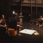 Eine Gruppe von Menschen sitzt in einem dunklen Theater- oder Proberaum im Kreis und hört einer stehenden Frau zu, die offenbar etwas erklärt. Auf dem Boden liegen einige Requisiten, während Stühle, schwarze Vorhänge und Bühnenbeleuchtung die Szene umgeben.