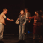 Auf einer dunkel beleuchteten Theaterbühne stehen mehrere junge Menschen in Kostümen und gestikulieren dramatisch miteinander. Im Mittelpunkt wirkt eine Frau in Jeansjacke verunsichert, während die anderen Figuren sie umringen und auf sie einreden.