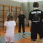In einer Turnhalle steht ein barfüßiger Mann in schwarzer Kleidung vor einer Gruppe und scheint etwas zu erklären oder anzuleiten. Mehrere Personen hören ihm zu, während an der Wand Holz-Sprossenwände und Sportgeräte zu sehen sind.