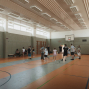 In einer großen Sporthalle stehen mehrere Jugendliche und Erwachsene auf dem Spielfeld, offenbar während eines Trainings oder Spiels. An der Wand hängt ein Basketballkorb, und durch die hohen Fenster fällt viel Licht in die Halle.