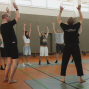 In einer Sporthalle stehen mehrere Personen im Kreis und heben gemeinsam die Arme, offenbar bei einer Übung oder einem Workshop. Rechts filmt ein Kameramann die Gruppe, während zwei Leiter im Vordergrund die Bewegung vormachen.