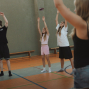 Mehrere Kinder und Jugendliche stehen in einer Sporthalle und heben bei einer Übung die Arme nach oben. Im Vordergrund ist eine Person unscharf zu sehen, während im Hintergrund Bänke und Hallenmarkierungen sichtbar sind.