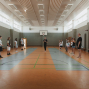 In einer hellen Sporthalle stehen mehrere Personen in zwei Reihen einander gegenüber, während eine Person in der Mitte offenbar etwas erklärt. Im Hintergrund sind ein Basketballkorb, Turnbänke und hohe Fenster zu sehen.