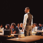 Ein Mann in Hemd und Weste steht in einem abgedunkelten Raum zwischen Reihen von Tischen, an denen viele junge Menschen sitzen. Die Szene wirkt wie eine Unterrichts- oder Theatervorstellung, mit Büchern und einer Tasche auf den Tischen im Vordergrund.