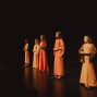 Fünf Mädchen stehen in langen, warmfarbenen Kostümen auf einer dunkel beleuchteten Theaterbühne. Links sind schemenhaft Zuschauer zu erkennen, während die Darstellerinnen ruhig mit gefalteten Händen nach vorn blicken.