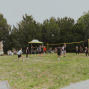 Mehrere Menschen spielen auf einer Wiese in einem Park Volleyball an einem gelben Netz. Im Hintergrund sitzen Zuschauer neben einem kleinen Pavillon, umgeben von Bäumen und Statuen.