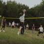Mehrere Jugendliche spielen auf einer Wiese im Freien Volleyball, während ein Spieler am Netz hochspringt und den Ball schlägt. Im Hintergrund stehen Zuschauer unter einem gelben Pavillon und zwischen hohen Bäumen.