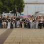 Eine große Gruppe junger Menschen steht mit Namensschildern auf einem gepflasterten Platz und posiert für ein Foto. Im Hintergrund sind Bäume, ein weiß verhülltes Gebäude mit Gerüsten sowie eine kuppelförmige Struktur zu sehen.