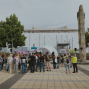 Eine Gruppe von Menschen versammelt sich auf einem gepflasterten Platz vor einer kleinen Bühne oder Kuppel, während im Hintergrund ein großes eingerüstetes Gebäude verhüllt ist. Rechts steht ein hohes steinernes Denkmal zwischen Bäumen, und die Szene wirkt wie eine öffentliche Veranstaltung im Freien.
