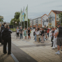 Eine Gruppe von Menschen versammelt sich auf einem städtischen Platz vor einem Gebäude, während links offenbar eine Ansprache oder Vorführung stattfindet. Im Hintergrund sind Fahnen, Bäume, Wohnhäuser und ein Bus zu sehen.