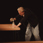 Eine Person mit blondem Haar steht an einem Holztisch in einer dunklen Umgebung und gießt Flüssigkeit aus einer kleinen Flasche in eine größere Flasche. Sie trägt einen schwarzen Hoodie und helle Jeans und wirkt dabei lächelnd und konzentriert.
