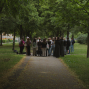 Eine Gruppe von Menschen steht auf einem gepflasterten Weg in einem grünen, von Bäumen gesäumten Park. Die Szene wirkt ruhig und schattig, während die Personen offenbar miteinander sprechen oder einer Führung zuhören.