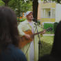 Ein Musiker in heller Kleidung und weißer Kappe singt im Freien in ein Mikrofon und spielt dabei Gitarre. Im Vordergrund sind unscharf die Köpfe von Zuschauern zu sehen, während im Hintergrund Bäume und Wohnhäuser stehen.