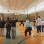 Eine Gruppe von Menschen steht in einer Sporthalle im Kreis und scheint gemeinsam eine Übung oder ein Spiel zu machen. Im Hintergrund sind ein Basketballkorb, Turngeräte und hohe Fenster zu sehen.