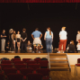 Eine Gruppe von Jugendlichen und Erwachsenen steht und sitzt auf einer Theaterbühne vor einem schwarzen Hintergrund und roten Vorhängen. Im Vordergrund sind leere rote Zuschauerstühle und eine Treppe zur Bühne zu sehen.