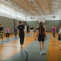 Eine Gruppe von Menschen steht in einer Sporthalle im Kreis, offenbar bei einer Tanz- oder Bewegungsübung. Die helle Halle hat markierte Spielfeldlinien, eine Basketballkorbwand und hohe Fenster.