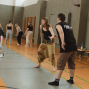 Mehrere Personen üben in einer Turnhalle gemeinsam Bewegungen oder Tanzschritte. Im Vordergrund stehen eine barfüßige Frau in einer gemusterten Hose und ein Mann im schwarzen ärmellosen Shirt einander gegenüber.