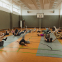 In einer Sporthalle liegen mehrere Personen verteilt auf dem Boden und führen eine Übung mit angehobenen Beinen und Armen aus. Im Hintergrund sind ein Basketballkorb, hohe Fenster und graue Hallenwände zu sehen.