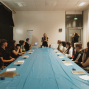 Eine Gruppe Jugendlicher sitzt in einem Seminarraum an einem langen Tisch mit blauer Abdeckung und schaut einer stehenden Kursleiterin zu. Auf dem Tisch liegen Papier, kleine Materialien und Geräte, während im Hintergrund ein Flipchart und weitere Personen zu sehen sind.