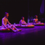 Drei Personen sitzen auf aufblasbaren Schlauchbooten auf einer dunklen Bühne, die in violettes Licht getaucht ist. Sie wirken wie Darsteller in einer Theater- oder Performance-Szene und schauen mit geöffneten Mündern nach vorne.