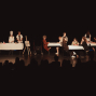 Auf einer dunklen Theaterbühne führen mehrere Kinder eine Szene mit gedeckten Tischen, Stühlen und Requisiten auf. Links steht ein Kind mit Kochmütze an einem Tisch, während rechts mehrere Kinder wie in einem Café oder Restaurant zusammensitzen.