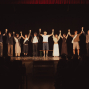 Eine Gruppe von Darstellerinnen und Darstellern steht auf einer Theaterbühne und hält sich nach einer Aufführung mit erhobenen Armen an den Händen. Vor ihnen sitzt das Publikum im Dunkeln, während die Bühne warm beleuchtet ist und ein roter Vorhang oben zu sehen ist.