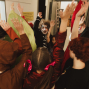 Eine Gruppe von verkleideten Kindern steht in einem Raum dicht beieinander und streckt die Arme nach oben. Einige tragen geschminkte Gesichter und bunte Kostüme, als würden sie feiern oder spielen.