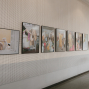 An einer perforierten weißen Galeriewand hängen mehrere gerahmte Fotografien in einer Reihe. Die Bilder zeigen überwiegend Mode- und Porträtszenen, während rechts ein heller Flur mit Tür zu sehen ist.