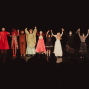 Eine Gruppe kostümierter Schauspieler steht auf einer dunklen Theaterbühne und hält sich nach einer Aufführung an den Händen. Vorne sind die Silhouetten des Publikums zu sehen, während die Darsteller gemeinsam Applaus entgegennehmen.