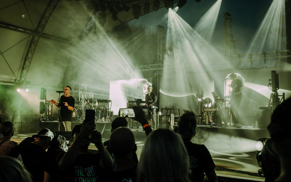 Eine Band spielt auf einer großen Bühne vor Publikum, während helle Scheinwerfer durch dichten Nebel strahlen. Im Vordergrund halten Zuschauer Smartphones hoch, um das Konzert zu filmen.