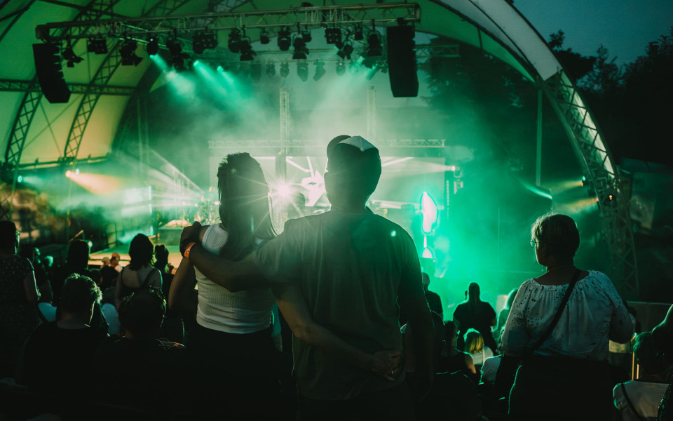 Ein Publikum schaut bei einem Open-Air-Konzert auf eine hell erleuchtete Bühne mit grünem Licht und Nebel. Im Vordergrund stehen zwei Personen eng umarmt als Silhouette vor der Bühne.